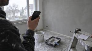 Maler hält ein Handy vor einer frisch verputzten Wand, am Boden Farbeimer, Farbrolle und Abdeckfolie