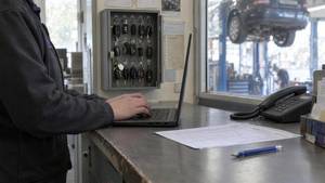 Hände tippen am Laptop auf dem Tresen einer Kfz-Werkstatt, daneben Telefon und Terminliste