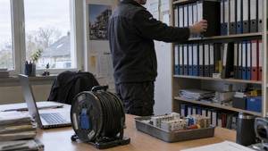 Büro eines kleinen Elektrobetriebs: ein Mitarbeiter zieht einen Ordner aus dem Regal, vorn Kabeltrommel, Laptop und Bauteile