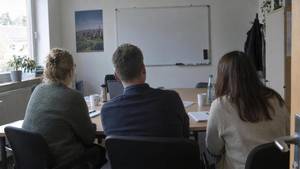 Drei Mitarbeitende von hinten an einem Besprechungstisch mit Laptop und Kaffeetassen, vorn ein leeres Whiteboard